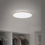 A round, modern ceiling light fixture is illuminated above an open closet with clothes on hangers. The room has light-colored walls and a window with horizontal blinds.