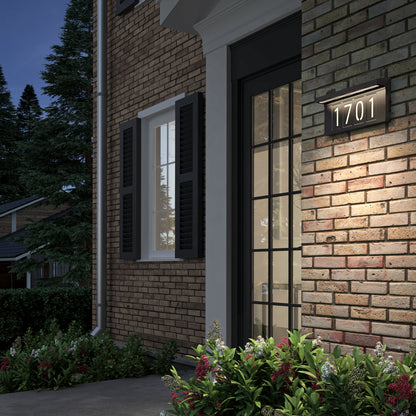 A brick house with black shutters features an Artika Bowen solar LED outdoor address wall light 5 CCT displaying 1701, a large glass door, front flower beds, and trees plus another house in the background.