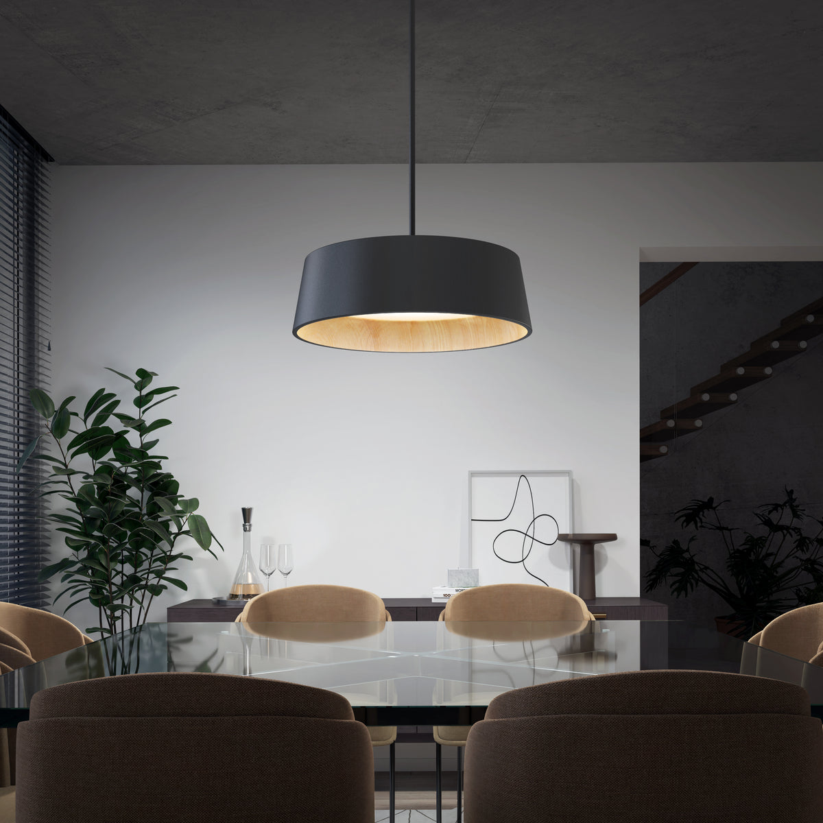 A modern dining room with a glass table, beige chairs, a large black pendant light, a potted plant, abstract artwork, and a staircase in the background. The room has minimalist decor and neutral colors.
