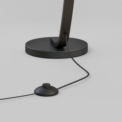 A close-up of the Artika Arc LED Floor Lamp base shows its power cord plugged in and a round foot switch, set against a light gray background.