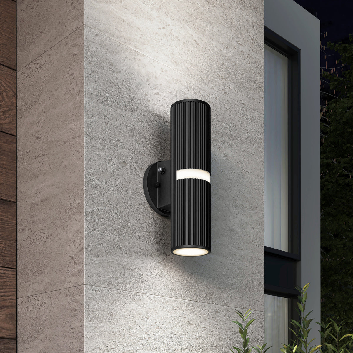 A modern black cylindrical outdoor wall light with horizontal grooves, mounted on a textured gray wall beside a window, illuminating upward and downward at night with plants visible below.