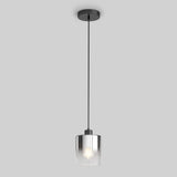 A modern pendant light with a black ceiling mount, a black cord, and a clear glass cylindrical shade, suspending a glowing light bulb against a plain gray background.