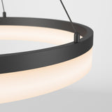 A close-up of a modern circular pendant light with a black frame and frosted white diffuser, suspended by two metal cables against a plain light background.