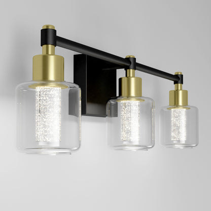 The Artika Champagne Globe bathroom vanity light features three clear glass shades, gold and black metal accents, and bright LED bulbs, shown mounted on a light gray wall.