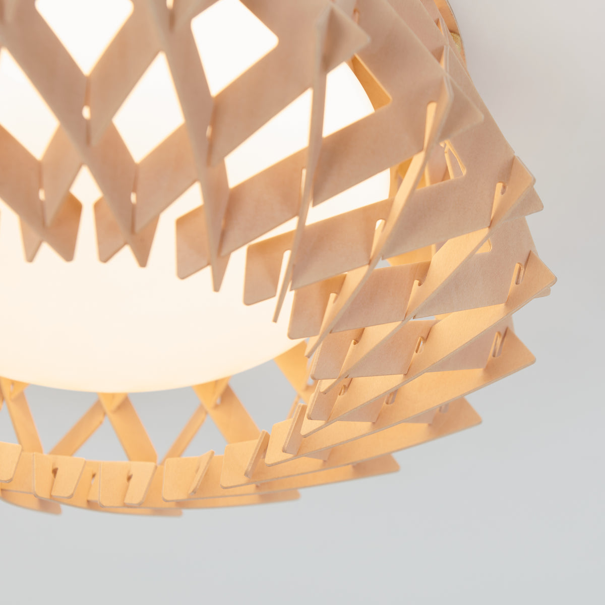A close-up of a modern ceiling light with a geometric wooden frame, featuring interlocking triangular shapes, surrounding a round white light bulb. The background is plain and light-colored.