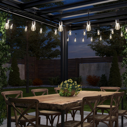 An Artika Blaze 20 LED Outdoor Solar String Lights set hangs above a wooden dining table with six chairs under a pergola. A potted plant decorates the table, while greenery and a wooden fence frame the patio.