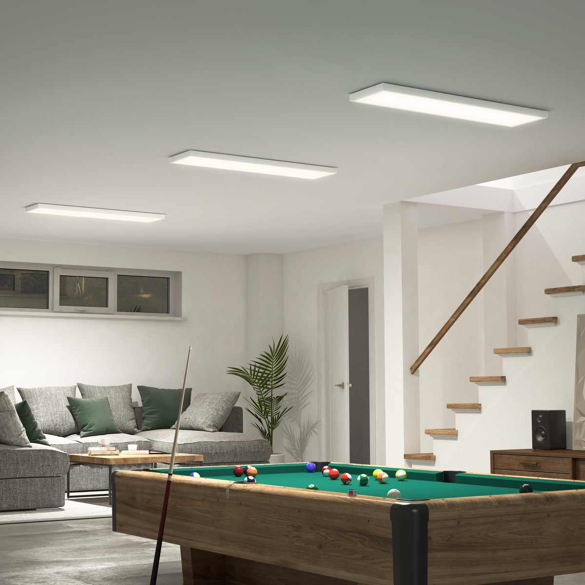 A modern basement room with a pool table in the foreground, a gray sectional sofa with green pillows, a coffee table, a potted plant, and a staircase leading upstairs. The space is bright and neatly organized.