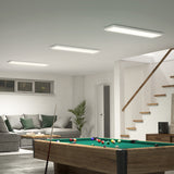 A modern basement room with a pool table in the foreground, a gray sectional sofa with green pillows, a coffee table, a potted plant, and a staircase leading upstairs. The space is bright and neatly organized.