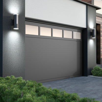 A modern home exterior features a large gray garage door, rectangular top windows, green shrubs in front, and two Artika Linea modern indoor-outdoor LED wall lights illuminating either side.