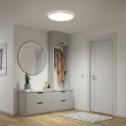A modern entryway features light wood flooring, gray cabinets and drawers, a round mirror, a potted plant, a hanging coat, and a white door illuminated by the Artika Jupiter 23 in. LED flush mount light with remote on the ceiling.