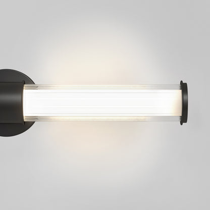 The Artika Kendra LED contemporary bathroom vanity light features a cylindrical clear glass shade, black metal end caps, and offers bright white light in 3 CCT options, mounted on a plain light gray wall.