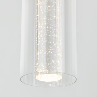 The Artika Bubble Flow 1-light pendant 2 features a modern cylindrical design with bubbles in the glass, casting a bright white glow against a plain light gray background.