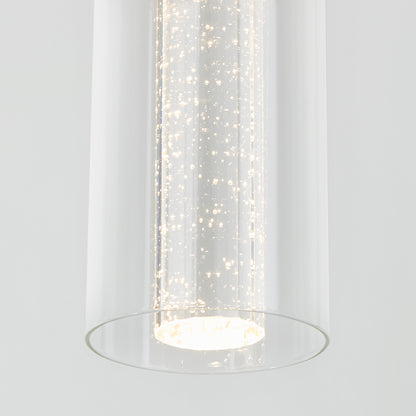 The Artika Bubble Flow 1-light pendant 2 features a modern cylindrical design with bubbles in the glass, casting a bright white glow against a plain light gray background.