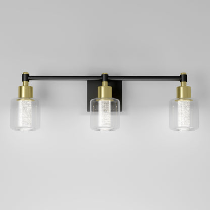 The Artika Champagne Globe bathroom vanity light is a modern wall-mounted fixture with three cylindrical glass shades, gold accents, and a black horizontal bar, shown against a plain white wall.