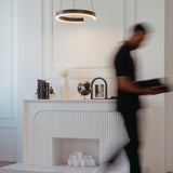 A blurred person walks past a modern white console decorated with books, a vase, a globe, framed art, and a cluster of candles below, under a circular pendant light on a white wall.