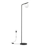 A modern black floor lamp with a round base and a slim, angled pole, featuring a spherical white lampshade at the top. The lamp is plugged into a wall outlet with a visible power cord and foot switch.