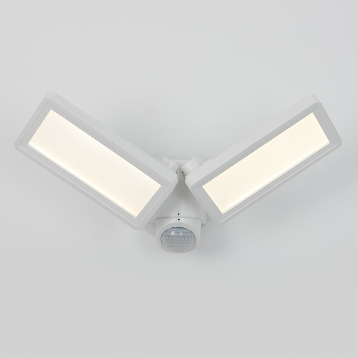 A white, modern LED security light fixture with two rectangular panels angled outward and a motion sensor centered below them, mounted on a plain, light gray background.