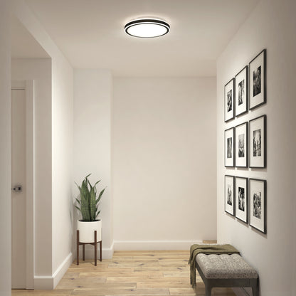 A minimal hallway with light wood floors, a potted plant on a stand, a bench with a green throw, a gallery wall of nine black and white framed photos, and the Artika Altitude 15 in. LED modern flush mount light 5 CCT.