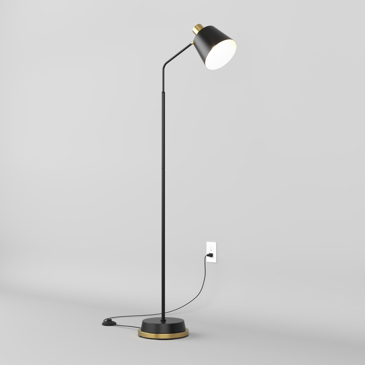 A modern black floor lamp with a gold accent stands on a light gray floor, plugged into a wall outlet. The lamp is turned on, casting light downward from its angled shade.