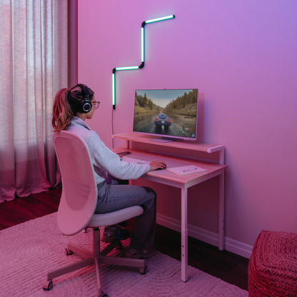 A person wearing headphones sits at a desk playing a car racing game on a monitor, with pink lighting from the Artika Fitch smart foldable RGB LED gaming floor lamp and modern decor featuring LED lights above the screen.
