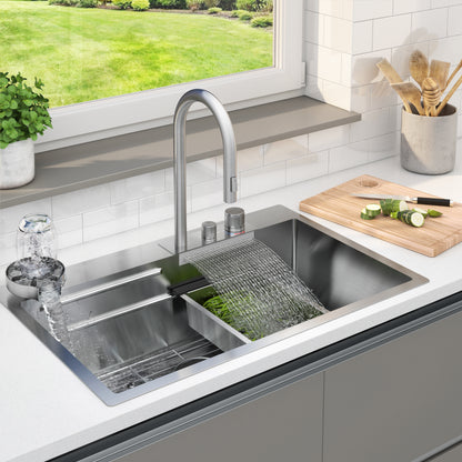 The Artika Neoflow single bowl stainless steel sink features a tall faucet running water into the basin as a glass fills on the left and chopped cucumbers rest on a cutting board to the right, with a window overlooking a green yard.