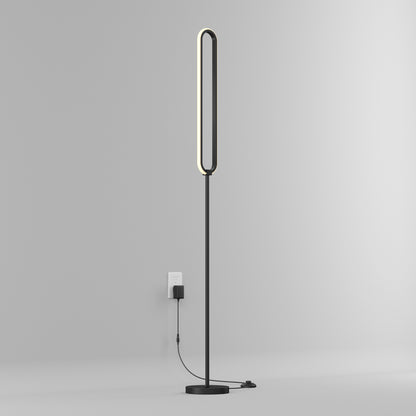 The Artika Leni dimmable LED modern floor lamp, featuring a sleek minimalist design and elongated oval light, stands against a plain gray wall and is plugged into a wall outlet.
