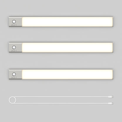Three Artika Outline under cabinet LED light 3CCT bars with silver ends and control buttons are arranged horizontally on a light gray background. Below them lies a white USB charging cable with connectors on both ends.