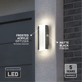 A modern outdoor wall light with a frosted acrylic diffuser and matte black finish is mounted beside a garage door. Labels highlight its features and a 5-year warranty. The light is turned on, illuminating the wall.