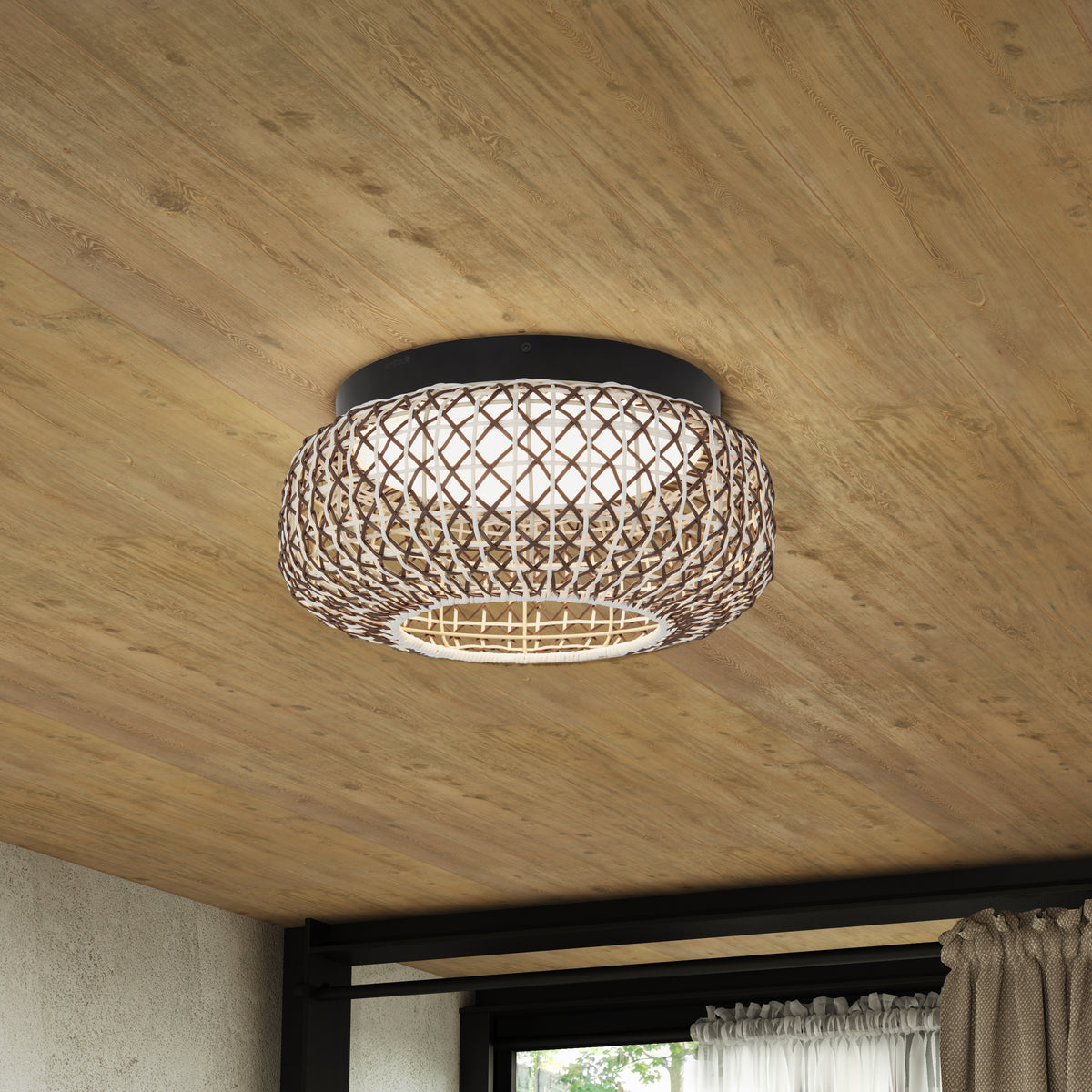 A woven rattan ceiling light fixture with a round, lattice-style design is mounted on a wooden ceiling. Daylight filters through a window with curtains in the background.
