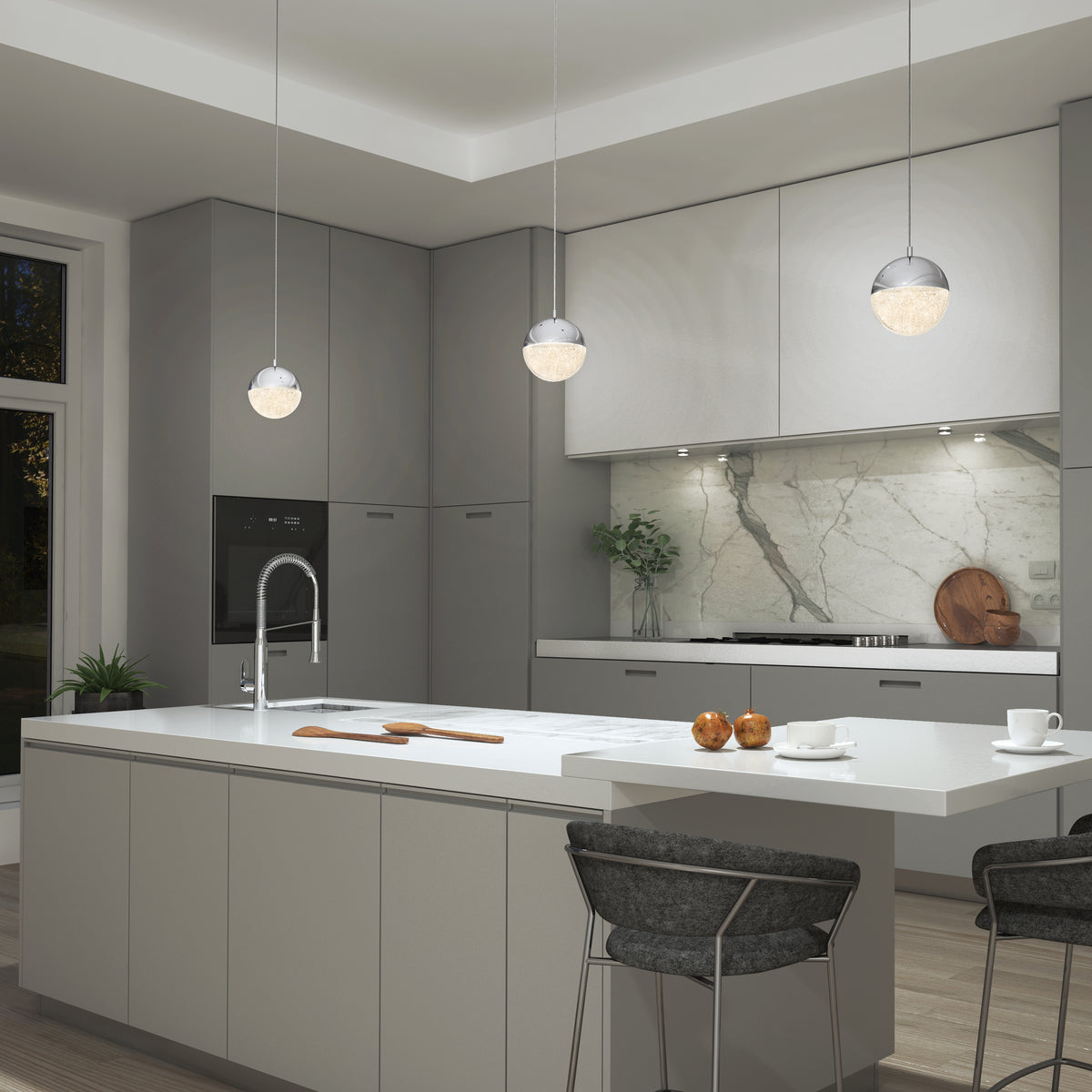 Modern kitchen with gray cabinets, a white island with sink, three spherical pendant lights, marble backsplash, and bar stools. Cutting boards and apples rest on the island; plants add greenery to the countertop.