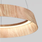 A close-up of a modern circular pendant light with a wood grain finish, suspended by thin wires against a plain light gray background. The light glows softly from inside the wooden ring.
