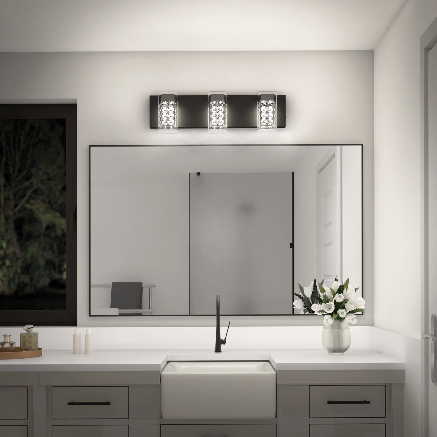A modern bathroom features Artika’s Crystal Cube LED bathroom vanity light (5 CCT) above a large mirror, with a white countertop, rectangular sink, black faucet, potted white flowers, and a window showing trees at night.