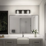 A modern bathroom with a large mirror, white countertop, rectangular sink, black faucet, a vase of white flowers, and a three-light fixture above the mirror. A window shows a dark, tree-filled view outside.