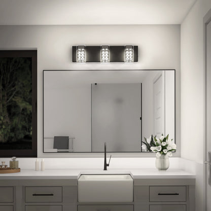 A modern bathroom features Artika’s Crystal Cube LED bathroom vanity light (5 CCT) above a large mirror, with a white countertop, rectangular sink, black faucet, potted white flowers, and a window showing trees at night.