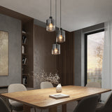 A modern dining room with a wooden table, four beige chairs, a vase with branches, and three pendant lights hanging above. Large window and built-in shelving are in the background, with neutral walls and natural light.