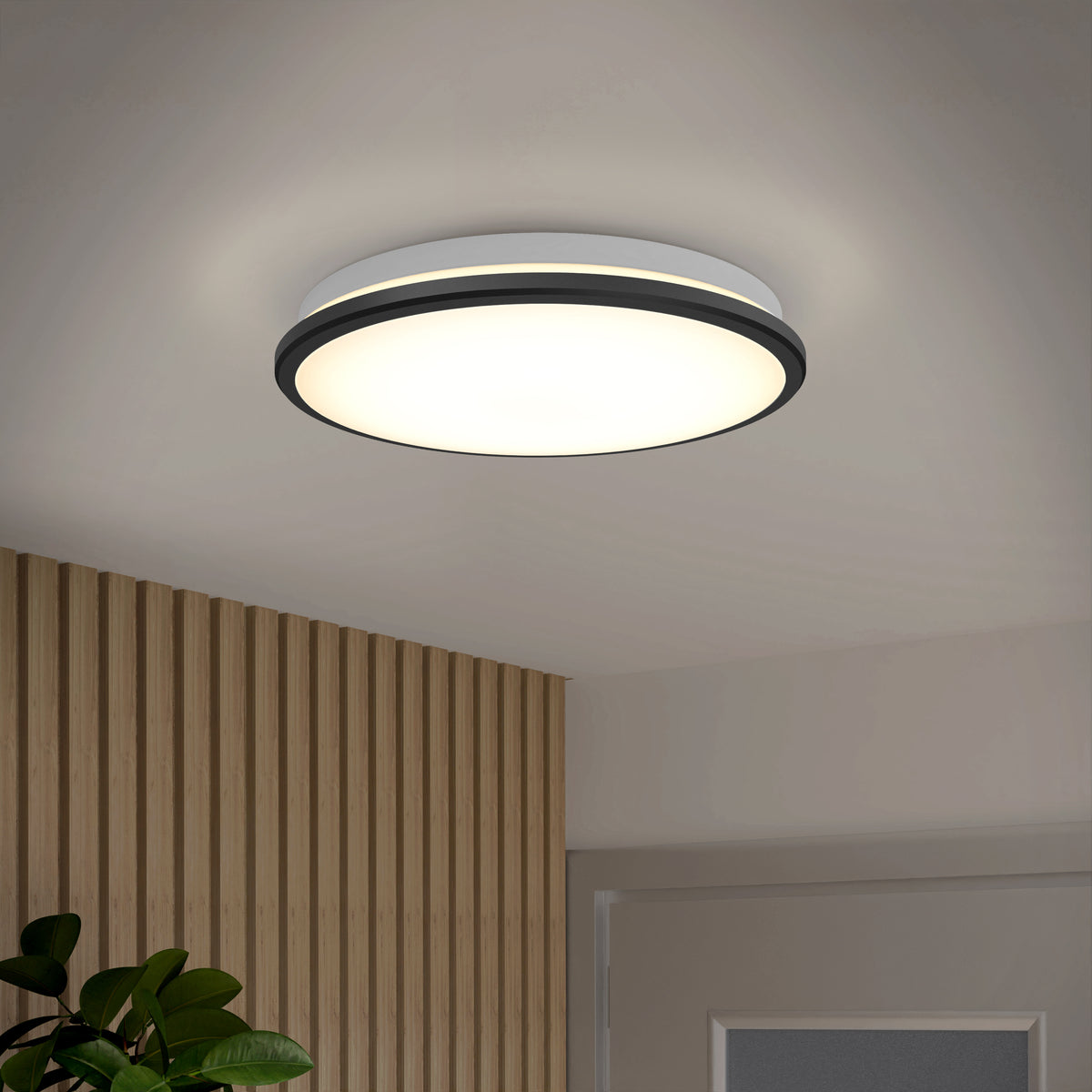 A modern, round ceiling light with a black trim is illuminated in a room with a wood-paneled wall, a green plant, and part of a white door visible.