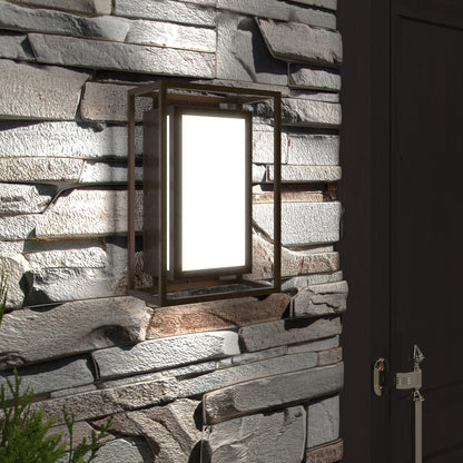 The Artika Ghost indoor-outdoor LED wall light with a metal frame is mounted on a textured stone wall beside a dark door, casting light and partially illuminating a green plant in the lower left corner.