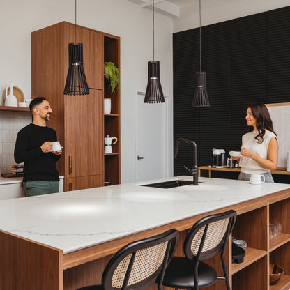 A man and woman stand on opposite sides of a modern kitchen island, each holding a mug and smiling. Wooden cabinets and a white marble countertop complement the Artika Kiyo modern LED pendant 3 CCT lights hanging above.