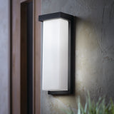 A modern rectangular outdoor wall sconce with a black frame and frosted glass is mounted on a textured gray wall near a doorway, with some green foliage visible at the bottom right.