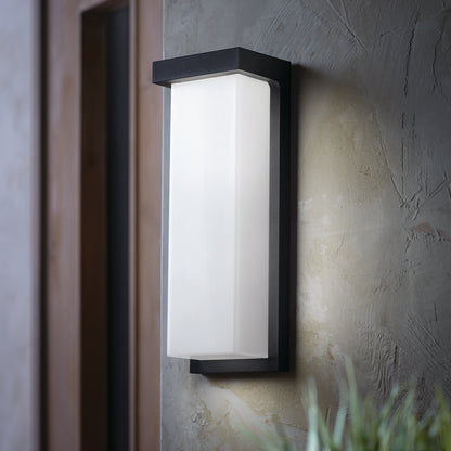 The Skeet LED indoor-outdoor light by Artika, featuring a black frame and frosted white glass, is mounted on a textured gray wall near a wooden door, with green plant leaves visible at the bottom right.