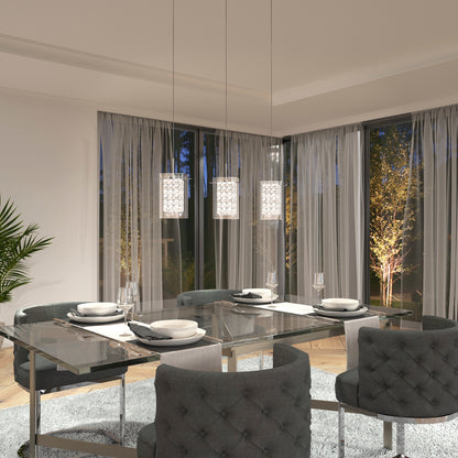 A modern dining room features a glass table set for four with gray upholstered chairs. Above, the Artika Crystal Cube 3-light LED pendant light shines, while large windows with sheer curtains reveal illuminated trees at night.