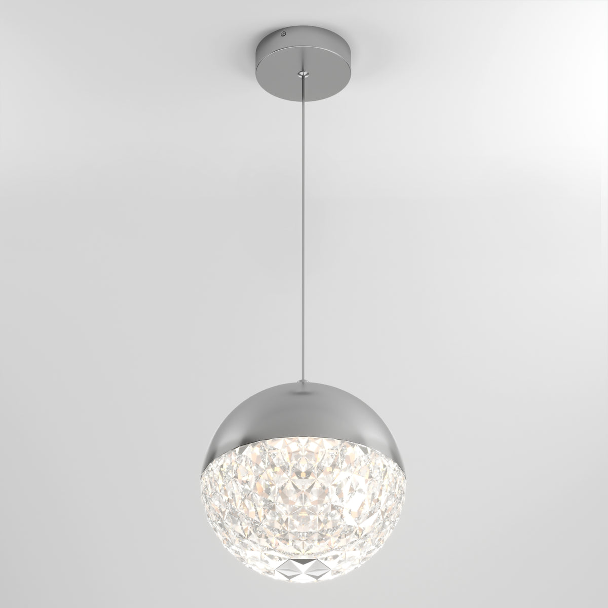The Artika Carat LED pendant light 3 CCT features a modern design with a round silver top, faceted crystal-like bottom, and hangs by a thin cord, illuminating elegantly against a plain white ceiling.