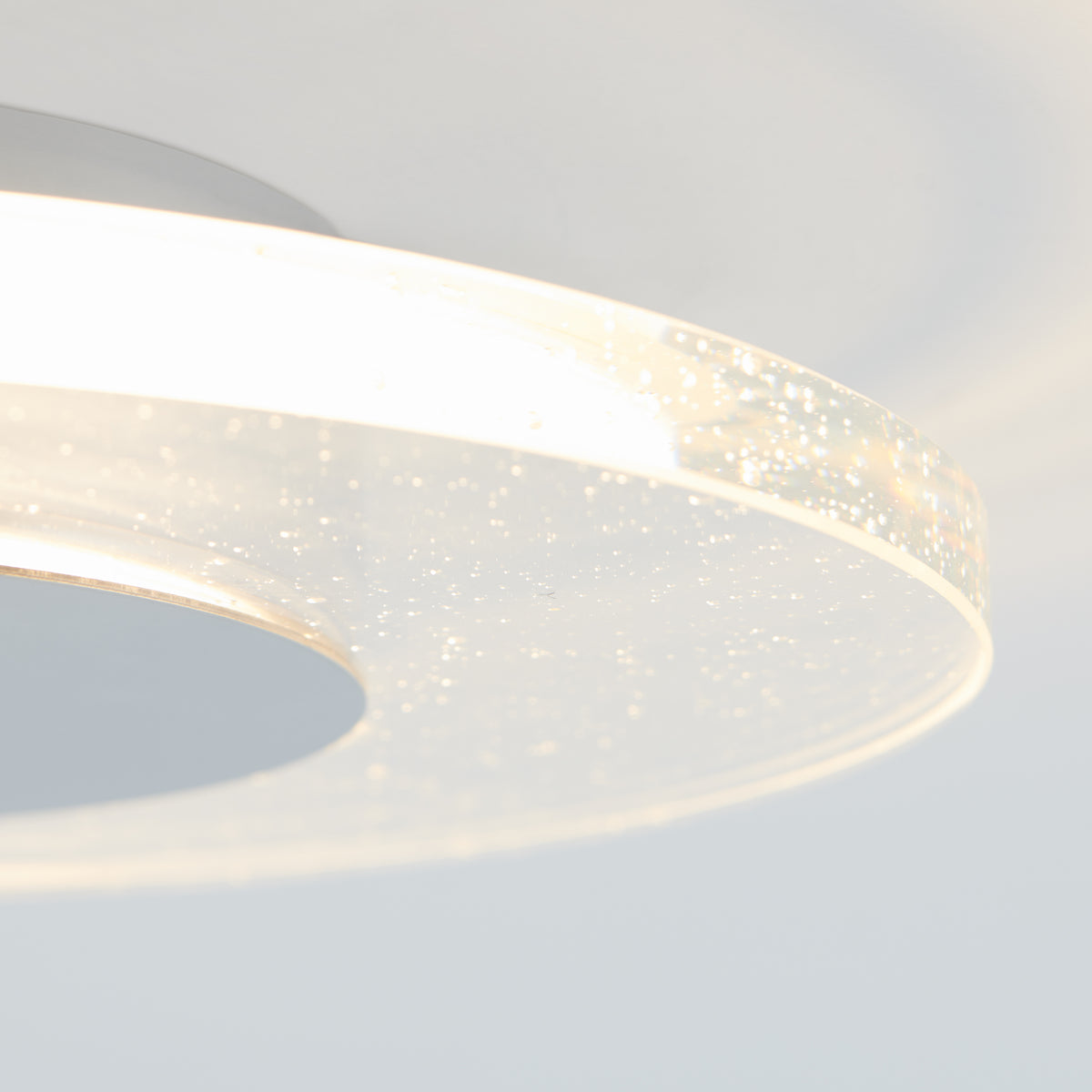 Close-up of a modern, circular ceiling light with a translucent edge embedded with small bubbles, emitting a soft, white glow against a light gray background.