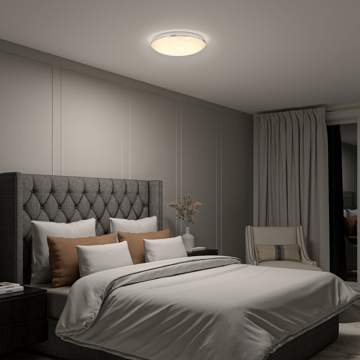 A modern bedroom with a tufted gray headboard, layered pillows, neutral bedding, a nightstand with a vase, a cushioned chair, floor-length curtains, and a round ceiling light illuminating the space.