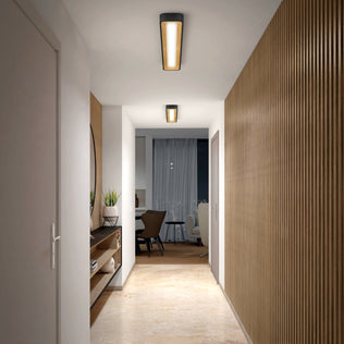 A modern hallway with wooden floors and slatted wood panels is lit by the Artika Alton 48 in. LED flush mount light. A side shelf holds plants and decor, leading to a dining area with dark chairs and a round mirror.