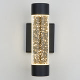 A modern wall sconce with a cylindrical glass body featuring tiny bubbles inside, illuminated and mounted on a square black base against a light-colored wall.