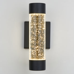 The Artika Bubble Element LED indoor-outdoor light features a cylindrical glass body filled with small bubbles, illuminated from both ends, and mounted on a black rectangular base against a light-colored wall.