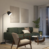 A modern living room with a green sofa, wooden armchair, floor lamp, glass coffee table, textured wall art, a potted plant, and large window with sheer curtains. The space has a cozy and minimalist style.