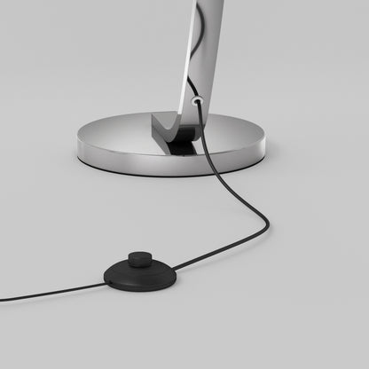 A close-up of the Artika Arc LED Floor Lamp’s chrome base with a power cord connected to a round black foot switch, resting on a smooth light gray surface.