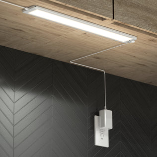 An Artika Maestro under cabinet LED light 3CCT is installed beneath a wooden cabinet, with its power cord plugged into a white outlet on a black chevron-patterned wall.